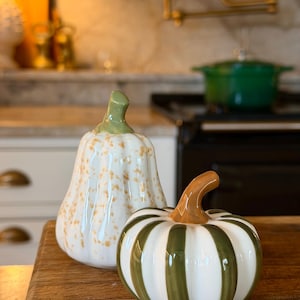 May include: Two ceramic pumpkin figurines. One is a tall, pear-shaped white pumpkin with a glossy finish and orange speckles, topped with a green stem. The other is a shorter, round white pumpkin with vertical olive green stripes and a brown stem.