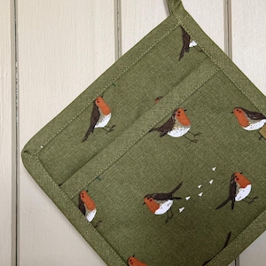 Peut inclure: Un manique carré en tissu de couleur vert olive avec un motif répétitif de petits rouges-gorges au poitrail orange et aux ailes brunes. La manique présente une bordure en tissu vert assorti et une petite boucle en tissu sur le coin supérieur.