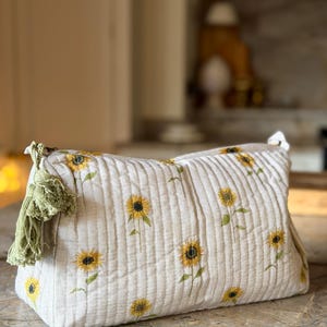 May include: A quilted beige pouch featuring a repeating sunflower print with yellow petals and dark centers. The rectangular bag has vertical stitching and a sage green tassel attached to the side zipper pull, providing a textured finish.