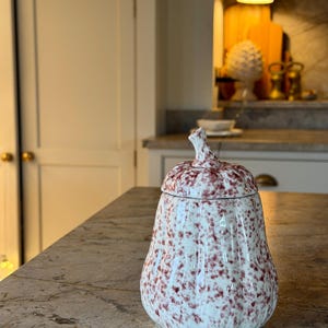 May include: A ceramic jar shaped like a pumpkin with a removable lid and a stem-shaped handle. The surface is white with a speckled pattern of reddish-brown spots. The jar features vertical ridges along its body, giving it a textured appearance.