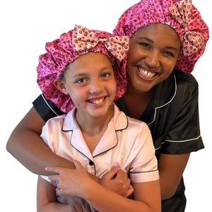 May include: Two matching hair bonnets in bright pink with a black heart pattern. Each bonnet features a gathered elastic edge and a decorative bow detail on the side. The fabric has a smooth, satin-like finish, providing a consistent look for both.