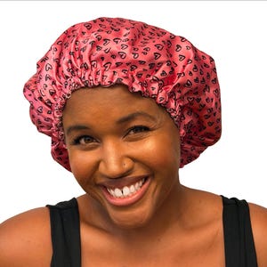 May include: A pink satin hair bonnet featuring a repeating pattern of small black heart outlines. The bonnet has a gathered elastic edge designed to fit securely around the head, providing a smooth and shiny texture across the fabric surface.