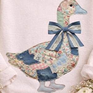 May include: A pale pink sweatshirt with a patchwork goose motif. The goose is crafted from floral, denim, and gingham fabrics, a blue bow with lace edging, and a blue gingham beak and feet. The design is centrally placed on the front of the garment.