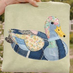 Peut inclure: Un sweat-shirt vert clair orné d'un appliqué de canard. Le canard est composé de différents patchworks de tissus, dont du denim, des imprimés floraux et du vichy. Il a un corps bleu, une tête fleurie et un bec jaune à pois noirs.