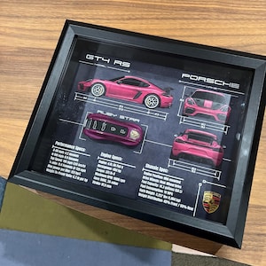 May include: Framed print featuring a pink Porsche GT4 RS, labeled "Ruby Star," with detailed diagrams and specifications. The print includes performance and engine specs, such as 0-60 mph in 3.2 seconds and a top speed of 196 mph. The Porsche logo is visible.