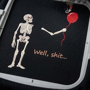 May include: A black fabric piece secured in an embroidery hoop featuring an embroidered design of a skeleton standing next to a detached skeletal arm holding a red balloon. The text "Well, shit..." is embroidered in brown thread below the arm.