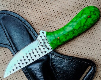 May include: A small decorative knife with a bright green handle and a silver blade. The handle has a marbled appearance and is secured with two small brass pins. The blade has triangular cutouts. The knife rests on a black leather sheath.