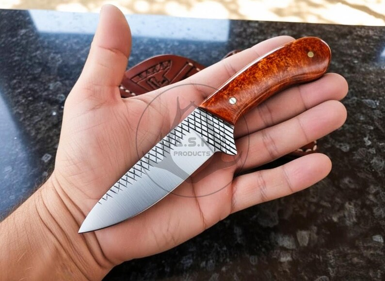 May include: A small, handcrafted knife with a polished, wooden handle and a silver blade. The handle is a warm, reddish-brown colour, and the blade has a textured pattern near the handle. The knife is held in a hand, showcasing its size and design.