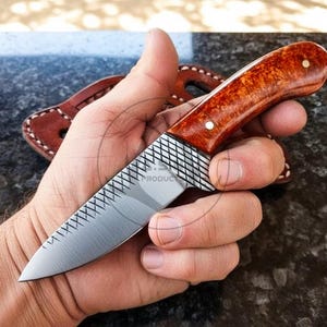 May include: A handcrafted knife with a wooden handle and a silver blade. The handle is a warm brown colour with a polished finish, and the blade has a textured pattern. A leather sheath is visible in the background.