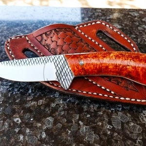 May include: A handcrafted knife with a polished, reddish-brown handle and a silver blade rests on a brown leather sheath. The sheath features decorative stitching and a textured pattern. The knife is a tool.