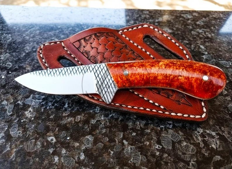 May include: A handcrafted knife with a polished, reddish-brown handle and a silver blade rests on a brown leather sheath. The sheath features decorative stitching and a textured pattern. The knife is a tool.
