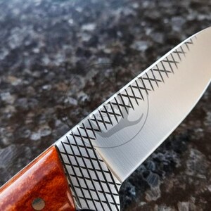 May include: Close-up of a stainless steel knife blade with a decorative etched pattern. The handle is a reddish-brown colour. The blade has a logo etched into the metal.