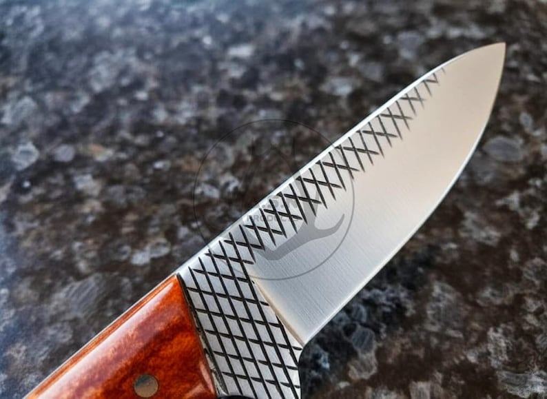 May include: Close-up of a stainless steel knife blade with a decorative etched pattern. The handle is a reddish-brown colour. The blade has a logo etched into the metal.
