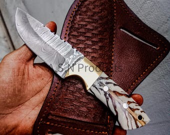 May include: A Damascus steel knife with a patterned blade and a handle featuring a white and brown design. The knife has a brass bolster and is paired with a brown leather sheath with a woven texture.