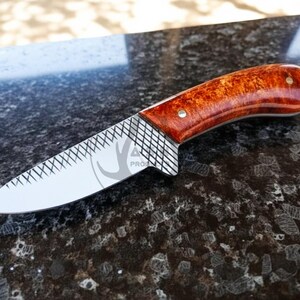 May include: A handcrafted knife with a polished blade and a reddish-brown wooden handle. The blade features a decorative pattern near the handle. The knife is designed for outdoor use and measures approximately 20 cm in length.