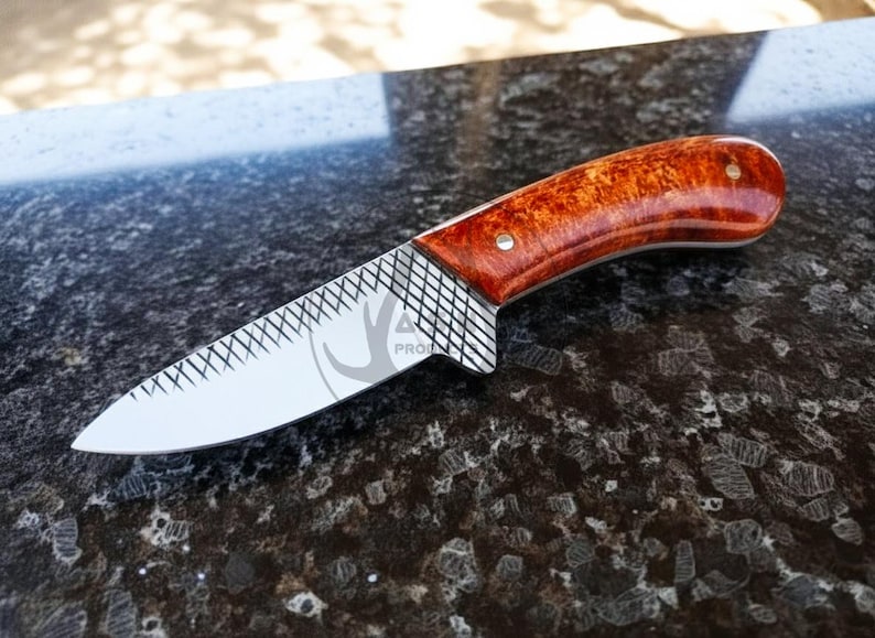 May include: A handcrafted knife with a polished blade and a reddish-brown wooden handle. The blade features a decorative pattern near the handle. The knife is designed for outdoor use and measures approximately 20 cm in length.