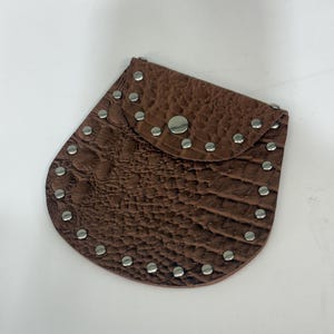 May include: A brown pouch with a textured, embossed surface resembling reptile skin. The pouch features a rounded flap closure secured by a single silver-toned metal snap button and is decorated with a border of small, round silver-toned metal studs.