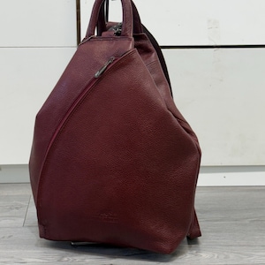 Könnte beinhalten: Ein tiefburgunderfarbener Rucksack aus strukturiertem Leder mit dreieckiger Silhouette, einem einzelnen Tragegriff und einer diagonalen Reißverschlusstasche auf der Vorderseite. Die Tasche verfügt über einen Hauptreißverschluss und verstellbare Riemen.