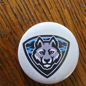 May include: A round, white button featuring a stylized wolf head within a shield design. The wolf is gray and black, set against a blue and black background. The button is on a wooden surface.