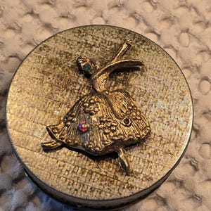 May include: A gold-colored round trinket box with a ballerina design on the lid. The ballerina is made of metal and has rhinestones for embellishments.