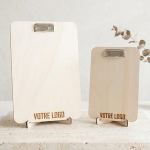 May include: Two light-colored wooden clipboards of different sizes, each featuring a silver metal clip at the top and a wooden stand at the base. The text "VOTRE LOGO" is engraved in a dark font at the bottom of each vertical rectangular board.