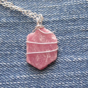 May include: A pink gemstone pendant wrapped in silver wire, hanging from a silver chain. The pendant is a natural, rough-cut stone with a unique shape.