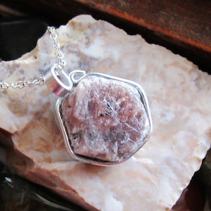 May include: A silver necklace featuring a raw, pink gemstone pendant. The pendant is a rough, irregularly shaped stone set in a silver bezel. The chain is a delicate silver link. The gemstone has a textured surface with variations in color.