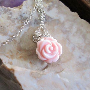 May include: A delicate silver chain necklace featuring a light pink rose pendant. The rose pendant is intricately detailed, showcasing a spiral design. The necklace is displayed on a textured, light-colored surface.