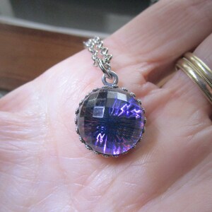 May include: A silver chain necklace with a round pendant featuring a purple, faceted glass stone set in a silver bezel.