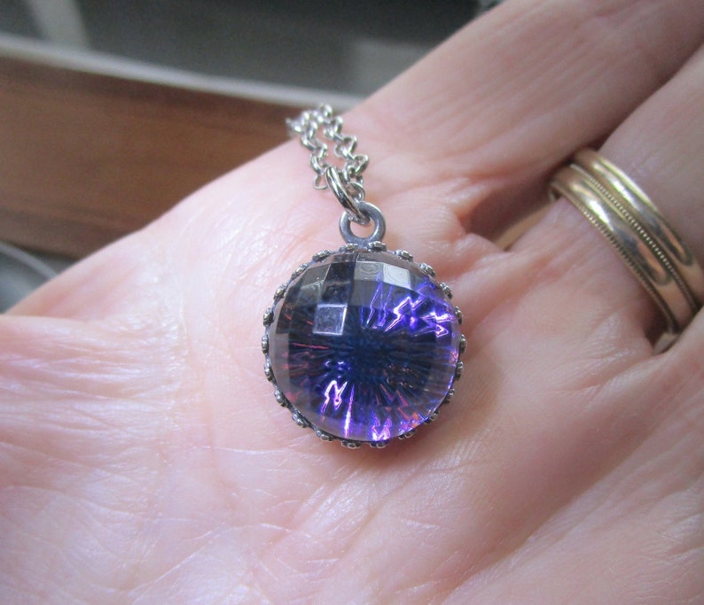May include: A silver chain necklace with a round pendant featuring a purple, faceted glass stone set in a silver bezel.