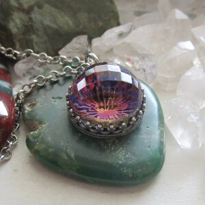 May include: A silver chain necklace with a pendant featuring a large, iridescent, faceted gemstone set in a silver bezel. The gemstone has a purple, red, and orange color scheme. The necklace is displayed on a green stone.