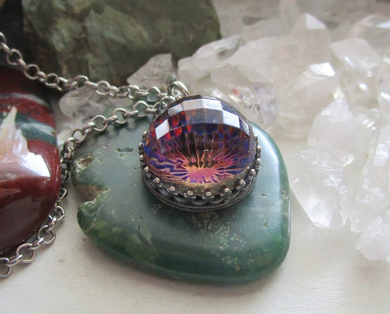 May include: A silver chain necklace with a pendant featuring a large, iridescent, faceted gemstone set in a silver bezel. The gemstone has a purple, red, and orange color scheme. The necklace is displayed on a green stone.
