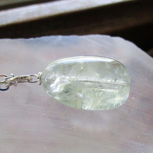 K&ouml;nnte beinhalten: Ein klarer, ovaler Anh&auml;nger mit einer silbernen Kette. Der Anh&auml;nger hat eine blassgr&uuml;ne Farbe und scheint aus einem durchscheinenden Stein zu bestehen. Die Kette ist eine filigrane Silber-Gliederkette. Der Schmuck wird auf einer hellen Oberfl&auml;che pr&auml;sentiert.