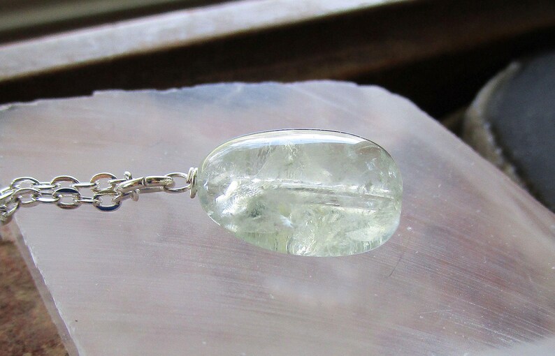 K&ouml;nnte beinhalten: Ein klarer, ovaler Anh&auml;nger mit einer silbernen Kette. Der Anh&auml;nger hat eine blassgr&uuml;ne Farbe und scheint aus einem durchscheinenden Stein zu bestehen. Die Kette ist eine filigrane Silber-Gliederkette. Der Schmuck wird auf einer hellen Oberfl&auml;che pr&auml;sentiert.