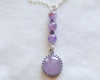 May include: A silver chain necklace with a pendant featuring a large, round, light purple gemstone set in a silver bezel with a decorative border. The necklace also features three smaller, round, light purple gemstones.
