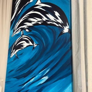 Op de afbeelding: Abstracte schildering van twee dolfijnen die uit een blauwe golf springen. De dolfijnen zijn zwart en wit, en de golf is diepblauw met witte highlights. Het schilderij is op canvas en heeft een witte rand.