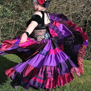 May include: A woman in a swirling, patchwork skirt in shades of purple, pink, and burgundy. The skirt is the focal point, with a variety of fabrics and patterns. She wears a black top and a floral hair accessory.