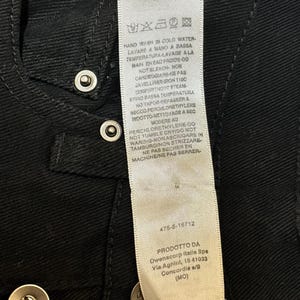 May include: Close-up of a black garment with silver snap closures. A white tag with care instructions and product information is visible. The tag includes text in multiple languages.