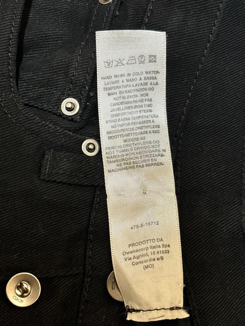 May include: Close-up of a black garment with silver snap closures. A white tag with care instructions and product information is visible. The tag includes text in multiple languages.