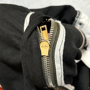 May include: Close-up of black denim fabric with a gold zipper and white stripes. The zipper is open, revealing the metal teeth and a gold pull tab with the brand name. The fabric has a textured appearance.