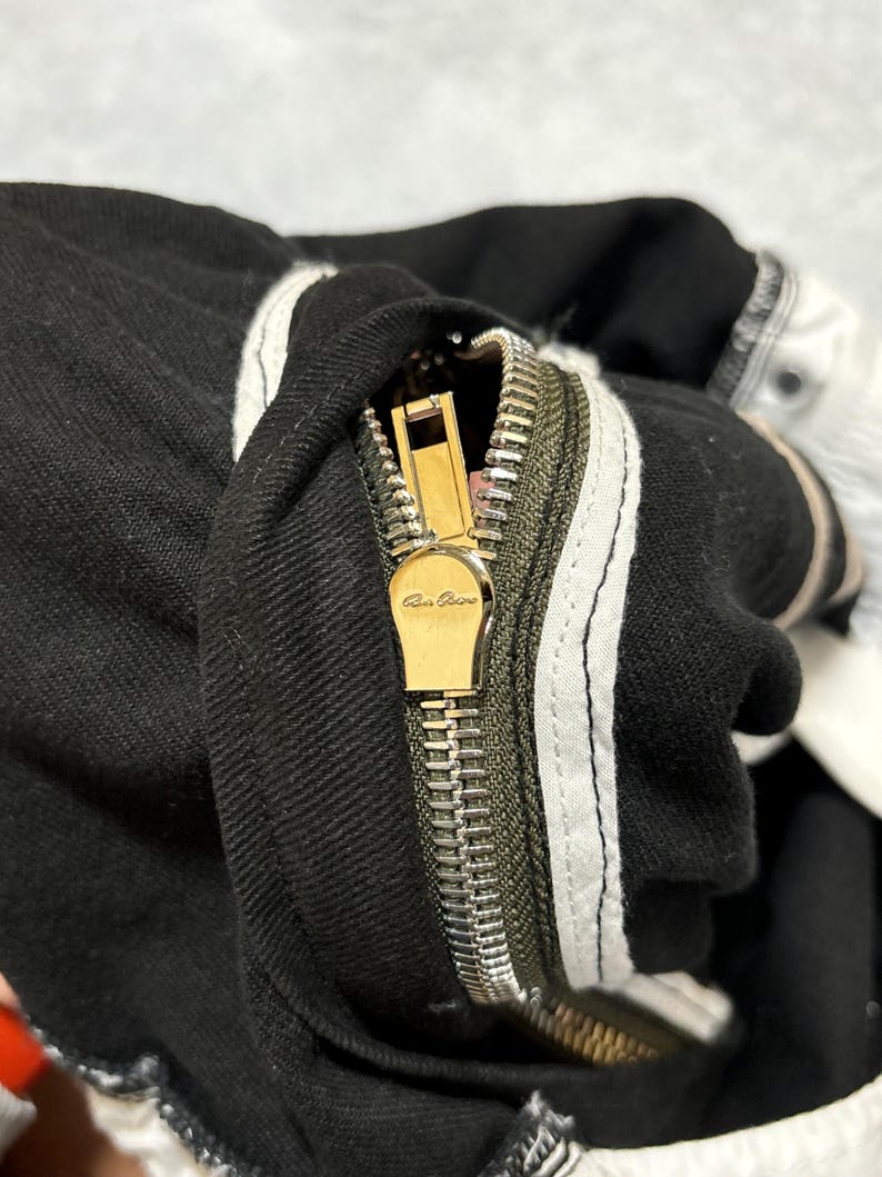 May include: Close-up of black denim fabric with a gold zipper and white stripes. The zipper is open, revealing the metal teeth and a gold pull tab with the brand name. The fabric has a textured appearance.