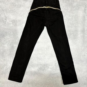 May include: Black corduroy pants with a gold-colored zipper that runs across the back. The pants have a high waist and a straight leg design. The zipper is a prominent design element.