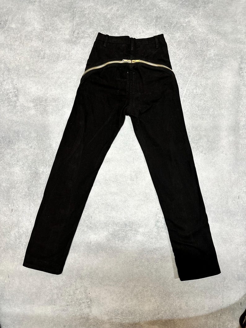 May include: Black corduroy pants with a gold-colored zipper that runs across the back. The pants have a high waist and a straight leg design. The zipper is a prominent design element.