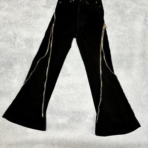 May include: Black flared pants with silver zippers running along the front of each leg. The pants have a high waist and a button and zipper closure. The fabric appears to be a soft, velvet-like material.