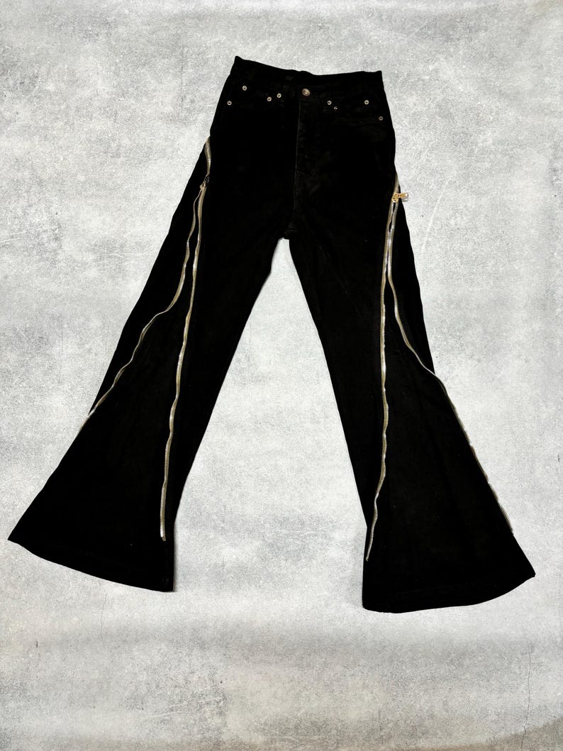 May include: Black flared pants with silver zippers running along the front of each leg. The pants have a high waist and a button and zipper closure. The fabric appears to be a soft, velvet-like material.