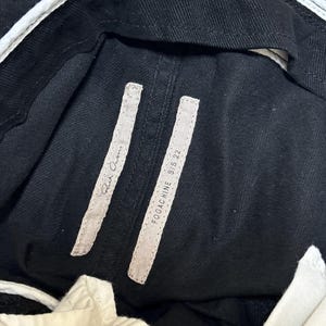 May include: Close-up of black denim pants with white accents. The pants have two rectangular tags with the text "Rick Owens" and "FOGACHINE S/S 22". The pants have a white waistband.