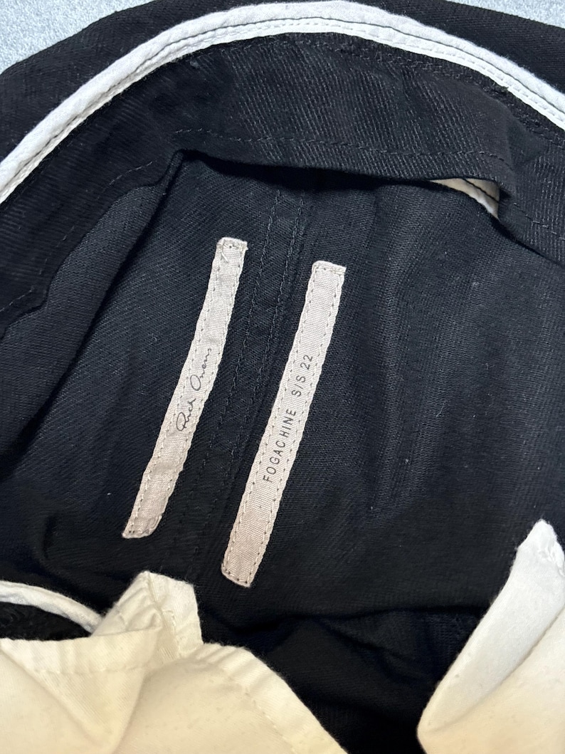 May include: Close-up of black denim pants with white accents. The pants have two rectangular tags with the text "Rick Owens" and "FOGACHINE S/S 22". The pants have a white waistband.