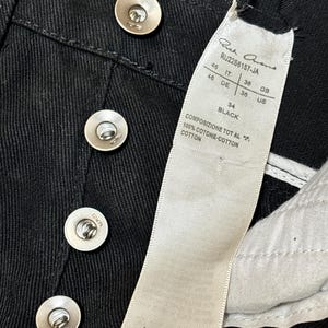 May include: Close-up of black denim pants with silver metal buttons. A white tag displays the brand name, style number, and size information. The tag also indicates the pants are made in Italy.