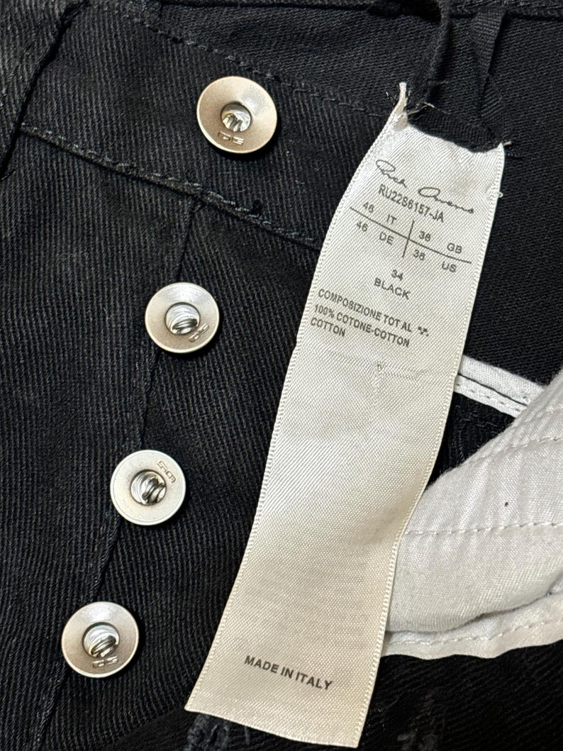 May include: Close-up of black denim pants with silver metal buttons. A white tag displays the brand name, style number, and size information. The tag also indicates the pants are made in Italy.