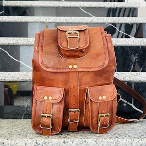 Puede incluir: Una mochila de cuero marrón con cierre de solapa y dos bolsillos delanteros. La mochila tiene correas ajustables y hebillas de color latón. La bolsa está sobre una superficie de mármol.